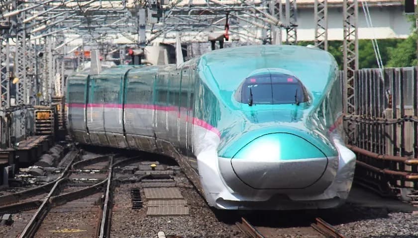An E5 series Shinkansen in green and white waiting at a platform, seen head-on along the track with overhead lines above it.